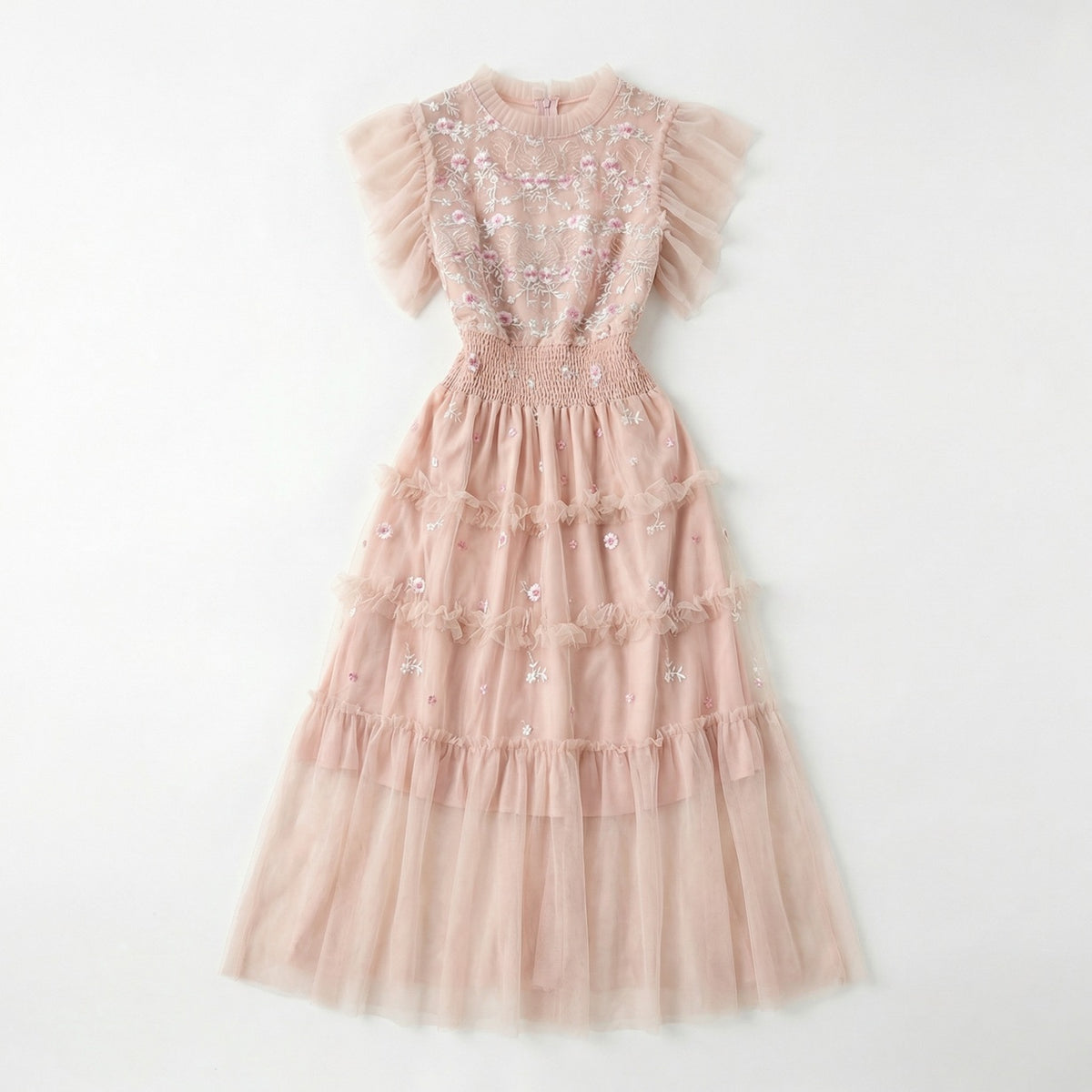 Full-length view of the Domenica pink mesh maxi dress featuring white and pink floral embroidery, a smocked waistline, and a tiered ruffled skirt laid out on a white faux fur rug.