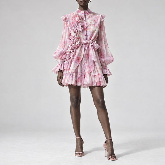 A model wearing a pink and white floral print long-sleeve mini dress with 3D floral appliqués and a matching fabric tie belt, paired with nude high-heeled sandals, against a light gray studio background.